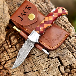 May include: A folding knife with a patterned blade and a handle made of wood squares in shades of red and brown. The knife rests on a brown leather sheath with the name "JACK" embossed on it. A small sharpening rod is also included.