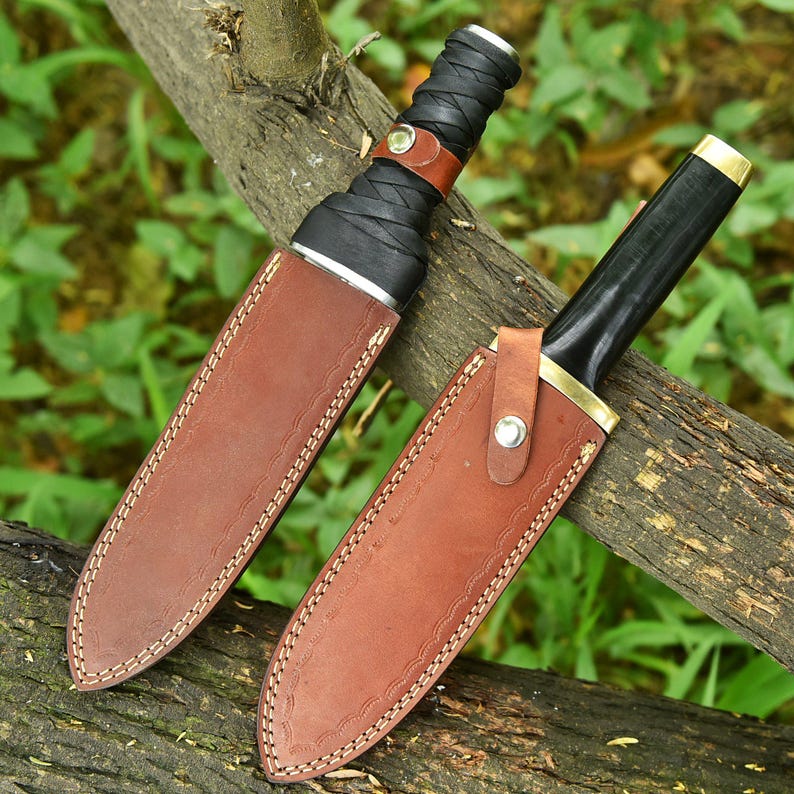 May include: Two hunting knives with brown leather sheaths. One knife has a black wrapped handle and a brown leather strap. The other knife has a black handle with a gold-coloured accent. Both knives are resting on a tree branch.