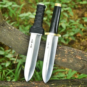May include: Two daggers with silver blades and black handles. One handle is wrapped in black leather, the other is smooth with a gold accent. The blades have arrows etched on them. The daggers are displayed on a tree branch.