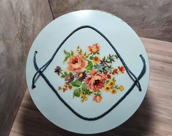 May include: A round, light blue decorative tin with a floral design. The flowers are in shades of pink, orange, yellow, and purple. A dark blue rope handle is attached to the tin. The tin is sitting on a wooden surface.