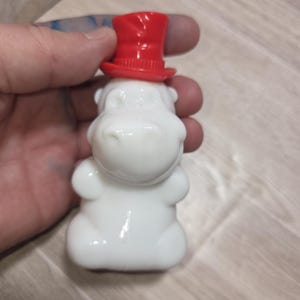 May include: A white ceramic hippo figurine wearing a bright red top hat. The hippo has a smooth, glossy finish and a whimsical design. The figurine is held in a hand.