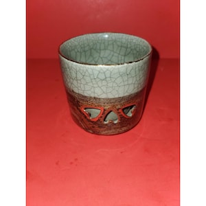 Puede incluir: Una taza de cerámica con acabado craquelado. La mitad superior es de color verde claro, y la mitad inferior es marrón con recortes en forma de corazón. La taza es cilíndrica y se encuentra sobre una superficie roja.