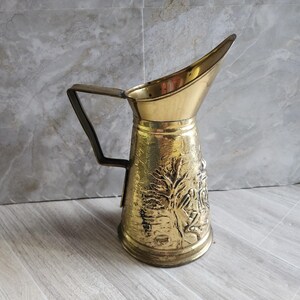 May include: A vintage brass pitcher with a handle and a spout. The pitcher has an embossed design of a tree and figures. The handle is a dark metal. The pitcher is approximately 8 inches tall.