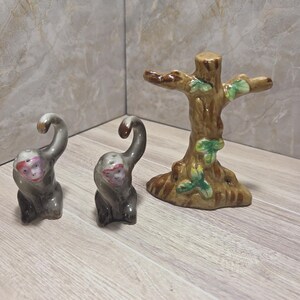May include: A ceramic salt and pepper shaker set featuring two monkey figures and a tree-shaped toothpick holder. The monkeys are grey with pink faces and brown accents. The tree is brown with green leaves.
