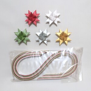 May include: A selection of paper star decorations in red, white, green, silver, and gold. Below the stars is a packet of paper strips in red, green, and gold, possibly for crafting. The items are set against a white backdrop.