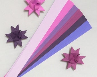 May include: A set of 12 origami paper strips in shades of pink, purple, and lavender. The strips are arranged in a fan shape, with the lightest shade at the top and the darkest shade at the bottom. Three origami stars made from the paper are also shown.