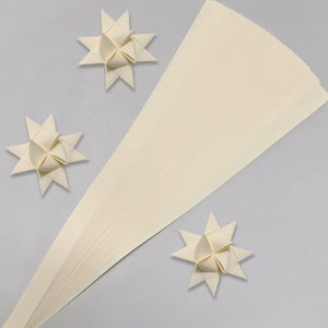 Könnte beinhalten: Eine Packung mit 50 elfenbeinfarbenen Papierstreifen zum Quilling. Die Streifen sind fächerförmig angeordnet und bilden ein Dreieck. Auf einem hellgrauen Hintergrund sind drei Origami-Sterne aus den Quillingstreifen zu sehen.