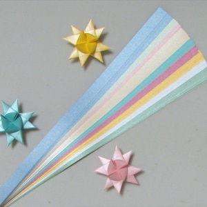 May include: An overhead shot shows colorful paper strips and origami stars. The paper strips are in pastel shades of blue, pink, yellow, and green. Three origami stars in yellow, teal, and pink are scattered around the paper strips.
