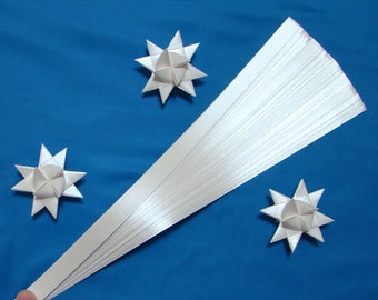 Puede incluir: Un paquete de tiras de papel de quilling blanco sobre un fondo azul. También se muestran tres estrellas de papel blanco.
