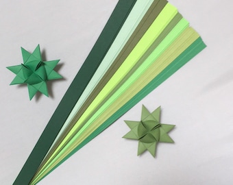 May include: A set of green, yellow, and white paper strips for quilling. The strips are arranged in a fan shape, with the darkest green at the bottom and the lightest white at the top. Two finished quilled stars are also shown.