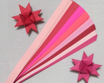 May include: A set of 50 paper strips in various shades of pink and red, perfect for origami. The strips are arranged in a fan shape, with the lightest shade at the top and the darkest shade at the bottom. Two origami stars made from the paper strips are also shown.