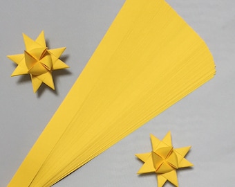 May include: A stack of yellow paper strips for making origami stars. Two finished origami stars are shown on a gray surface.