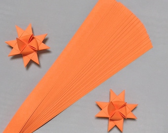 May include: Orange paper strips and two origami star ornaments on a gray background. The paper strips are fanned out, and the origami stars have pointed edges. The image is well-lit, showcasing the vibrant orange color.
