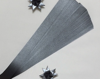 Puede incluir: Un paquete de 50 tiras de papel de origami plateado. Las tiras están dispuestas en forma de abanico. Se muestran dos estrellas de origami plateadas en la imagen.