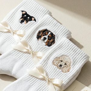 Könnte beinhalten: Drei Paar weiße Rippsocken, jeweils mit einem kleinen gestickten Hundeporträt. Die Socken sind mit einer cremefarbenen Satinschleife zusammengebunden. Eine kleine rechteckige Karte trägt den Text The Pet Memorial COLLECTION.