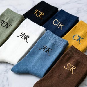 Puede incluir: Una colección de calcetines doblados en varios colores, incluyendo blanco, azul y marrón. Cada par presenta iniciales bordadas en un color contrastante. Los calcetines están cuidadosamente dispuestos, mostrando los diseños personalizados.