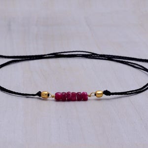 May include: A black string bracelet featuring a row of small, vibrant red gemstones. The bracelet has gold-colored beads at each end and is adjustable. The gemstones are a deep red color, and the bracelet is displayed on a light-colored surface.