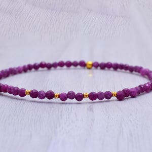 Puede incluir: Una delicada pulsera con pequeñas cuentas facetadas de color magenta intenso. La pulsera está adornada con pequeñas cuentas doradas. La joya se exhibe sobre una superficie clara y texturizada.