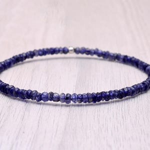 May include: A delicate bracelet composed of small, faceted, deep blue gemstones. The beads are strung together, forming a continuous loop. The bracelet is displayed on a light-colored, textured surface, showcasing its color and design.
