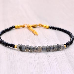 May include: A delicate bracelet with a central row of grey labradorite beads, flanked by black beads. The bracelet has gold-coloured accents and a clasp. The jewellery is displayed on a light-coloured wooden surface.