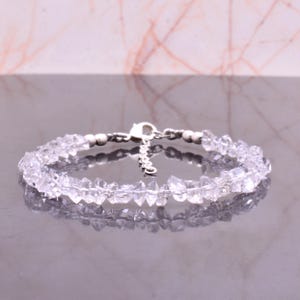 Peut inclure: Un bracelet en quartz clair avec un fermoir argenté. Le bracelet est composé de petites pierres de quartz de forme irrégulière. Le fermoir et la chaîne en argent sont visibles. Le bracelet est présenté sur une surface réfléchissante.