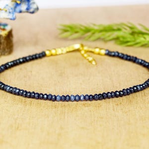 May include: A delicate dark blue beaded bracelet with a gold-colored clasp. The bracelet is composed of small, faceted beads, and the clasp is a simple hook and chain design. The bracelet is displayed on a light brown surface.