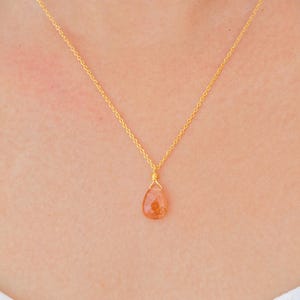 Può includere: Una collana color oro con un pendente arancione a forma di goccia. Il pendente è avvolto con filo metallico e appeso a una catena sottile. La collana è indossata intorno al collo.