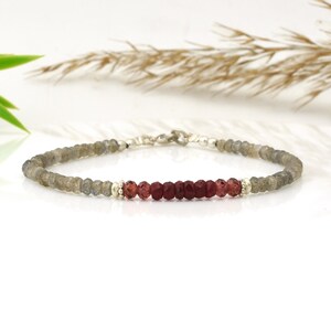 May include: A beaded bracelet featuring a combination of gray and red gemstones. The bracelet has a silver clasp and small silver beads separating the gray and red stones. The bracelet is displayed on a white surface.