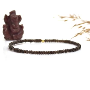 Könnte beinhalten: Ein zartes braunes Perlenarmband mit einem goldenen Verschluss. Im Hintergrund befindet sich eine kleine, geschnitzte braune Statue einer Gottheit. Das Armband besteht aus kleinen, facettierten Perlen.