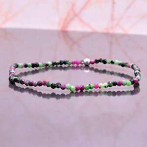 May include: A delicate bracelet composed of small, faceted beads in shades of green, black, and magenta. The beads are strung together, with a small silver-colored clasp. The bracelet is displayed on a reflective surface.