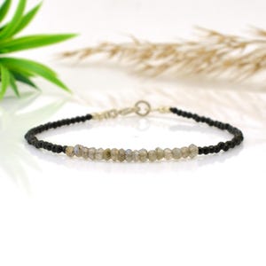 Puede incluir: Una delicada pulsera con una fila central de cuentas claras facetadas, flanqueada por cuentas negras. La pulsera está ensartada en una cadena y cierre plateados. La joya se exhibe sobre una superficie blanca con vegetación de fondo.