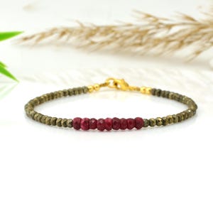 May include: A bracelet with olive-green and ruby-red gemstones. The bracelet has a gold-coloured clasp and connecting beads. The gemstones are small and faceted, creating a sparkling effect. The bracelet is displayed on a white surface.