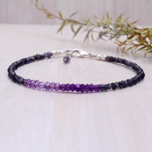 Può includere: Un delicato braccialetto con una sfumatura di pietre preziose viola, che vanno dal lavanda chiaro all'indaco scuro. Le perline sono infilate su una catena d'argento con un piccolo ciondolo e chiusura. Un gioiello.