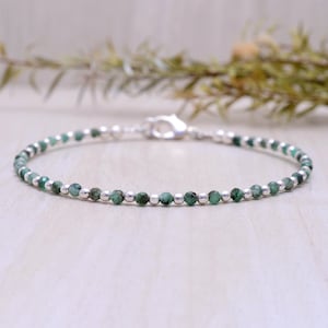 Könnte beinhalten: Ein zartes Armband mit kleinen, runden, smaragdgrünen Perlen, die mit silberfarbenen Perlen durchsetzt sind. Das Armband wird mit einem silberfarbenen Verschluss gesichert. Die Perlen sind aufgefädelt und bilden ein schlichtes, aber elegantes Design.