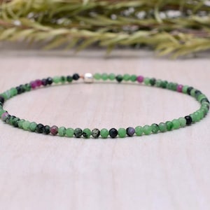 May include: A delicate bracelet made of small, round beads in shades of green, black, and a few pinkish-red accents. The beads are strung together, forming a flexible band. The bracelet is displayed on a light-coloured surface.