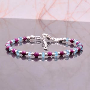 Puede incluir: Una pulsera de cuentas con piedras preciosas moradas y azul claro alternadas, intercaladas con pequeñas cuentas plateadas. La pulsera tiene un cierre plateado y un pequeño dije. Las piedras son redondas y espaciadas.