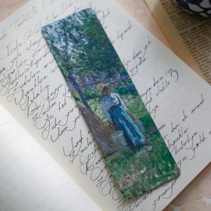 Pu&ograve; includere: Un segnalibro rettangolare con un dipinto impressionista di una persona con un vestito blu in un giardino. Il segnalibro &egrave; posto su una pagina di testo manoscritto, con uno sfondo bianco sporco.