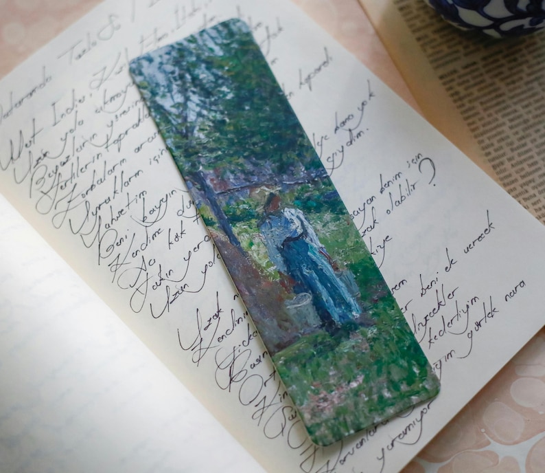 Pu&ograve; includere: Un segnalibro rettangolare con un dipinto impressionista di una persona con un vestito blu in un giardino. Il segnalibro &egrave; posto su una pagina di testo manoscritto, con uno sfondo bianco sporco.