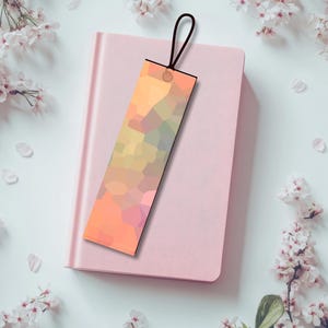 Pu&ograve; includere: Un segnalibro geometrico nei toni dell'arancione, giallo e rosa, con un cordino nero. Poggia su un quaderno rosa chiuso, circondato da fiori e petali bianchi.
