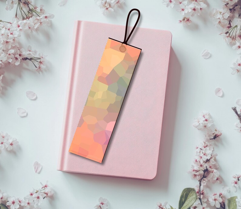 Pu&ograve; includere: Un segnalibro geometrico nei toni dell'arancione, giallo e rosa, con un cordino nero. Poggia su un quaderno rosa chiuso, circondato da fiori e petali bianchi.
