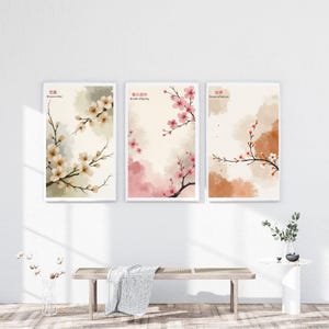 K&ouml;nnte beinhalten: Drei gerahmte Kunstdrucke mit Aquarellillustrationen von Kirschbl&uuml;tenzweigen. Die Drucke haben einen wei&szlig;en Rand und sind vor einem neutralen Hintergrund mit sanften Farbt&ouml;nen in Rosa, Beige und Braun platziert. Japanischer Text ist sichtbar.