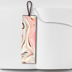 Pu&ograve; includere: Un segnalibro rettangolare con un design marmorizzato rosa, bianco e marrone. Il segnalibro ha un bordo nero e un anello marrone scuro nella parte superiore. Il segnalibro &egrave; posto all'interno di un quaderno bianco aperto.