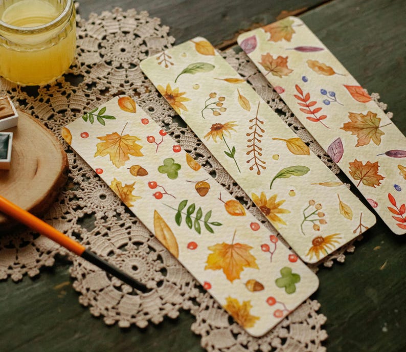 Pu&ograve; includere: Quattro segnalibri ad acquerello con disegni di foglie autunnali e fiori. I segnalibri presentano illustrazioni di foglie autunnali, ghiande, bacche e fiori nei toni del giallo, arancione e verde su sfondo crema. I segnalibri sono disposti su una tovaglietta di pizzo decorativa.