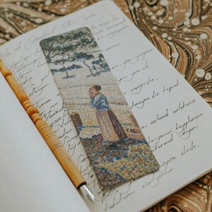 Pu&ograve; includere: Segnalibro decorativo con un dipinto di una donna in un campo, con alberi sullo sfondo. Il segnalibro &egrave; posto in un quaderno aperto con testo manoscritto e una penna di legno.