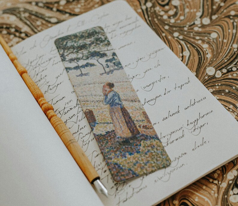 Pu&ograve; includere: Segnalibro decorativo con un dipinto di una donna in un campo, con alberi sullo sfondo. Il segnalibro &egrave; posto in un quaderno aperto con testo manoscritto e una penna di legno.