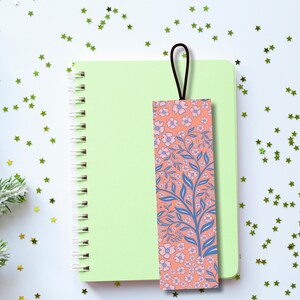 Pu&ograve; includere: Un segnalibro floreale con un anello marrone poggia su un taccuino a spirale verde chiaro. Il segnalibro presenta uno sfondo corallo con rami blu e fiori rosa. Il taccuino &egrave; su una superficie bianca cosparsa di coriandoli verdi a forma di stella.