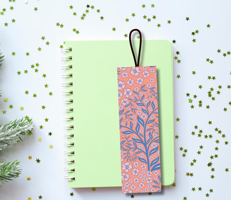 Pu&ograve; includere: Un segnalibro floreale con un anello marrone poggia su un taccuino a spirale verde chiaro. Il segnalibro presenta uno sfondo corallo con rami blu e fiori rosa. Il taccuino &egrave; su una superficie bianca cosparsa di coriandoli verdi a forma di stella.