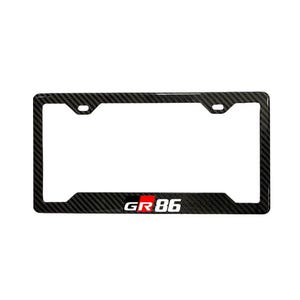 May include: Black carbon fiber license plate frame with the text "GR 86" in white and red. The frame has a rectangular shape with mounting holes at the top.