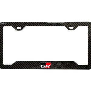 Könnte beinhalten: Ein schwarzer Kennzeichenrahmen mit einem Karbonfasermuster. Der Rahmen hat die Buchstaben "GR" in Weiß mit rotem Hintergrund. Der Rahmen hat vier Befestigungslöcher und eine rechteckige Form.
