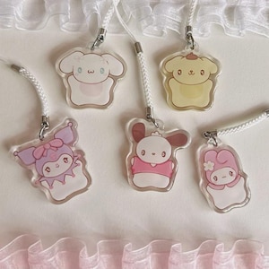 May include: Five clear acrylic charms with cartoon character designs. Each charm has a white cord for attaching to a phone or other device. The characters include a white dog, a yellow dog, a purple character with a bow, a brown bunny, and a pink character with a flower.
